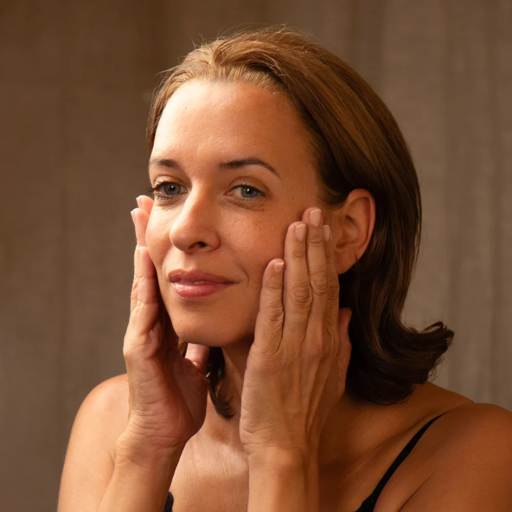 Woman gently applying LE PURE serum to her face, in warm natural light with a soft beige background.