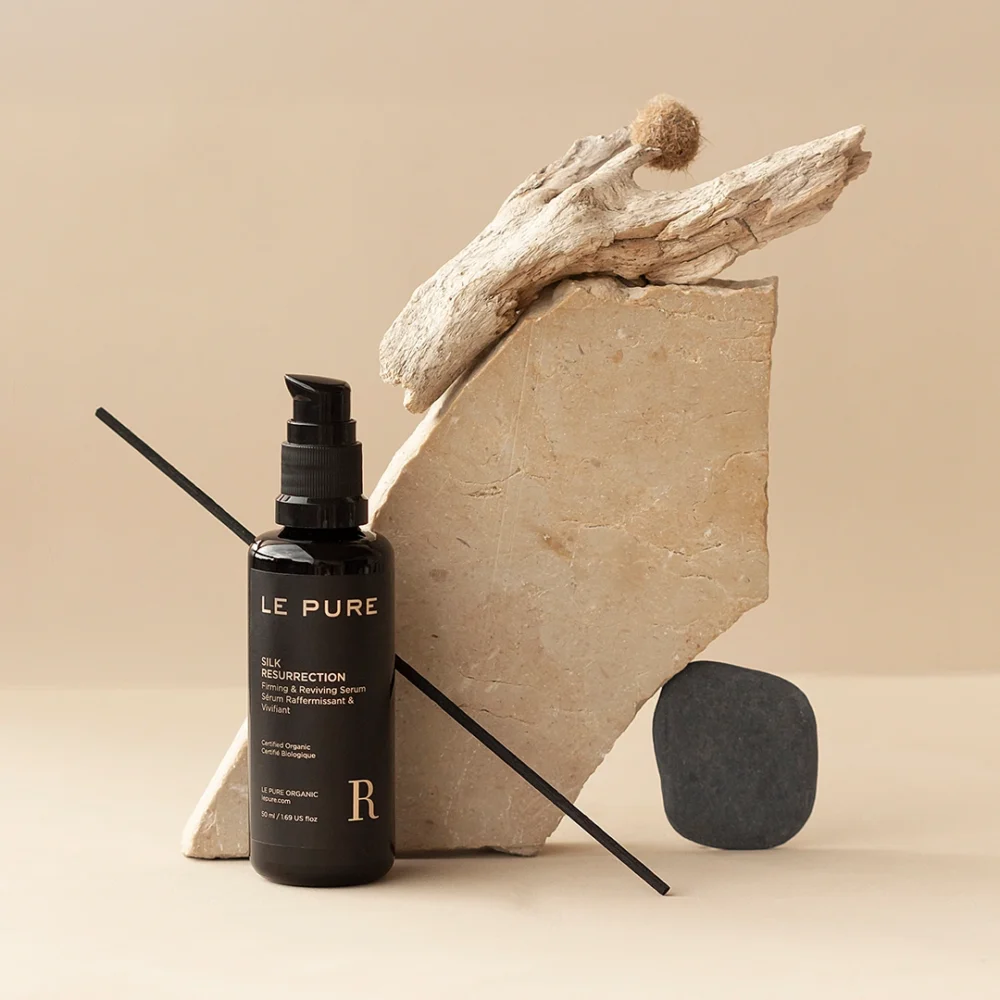 LE PURE Silk Resurrection phyto-gel serum in violet glass bottle, styled with natural stone and driftwood on a beige background.